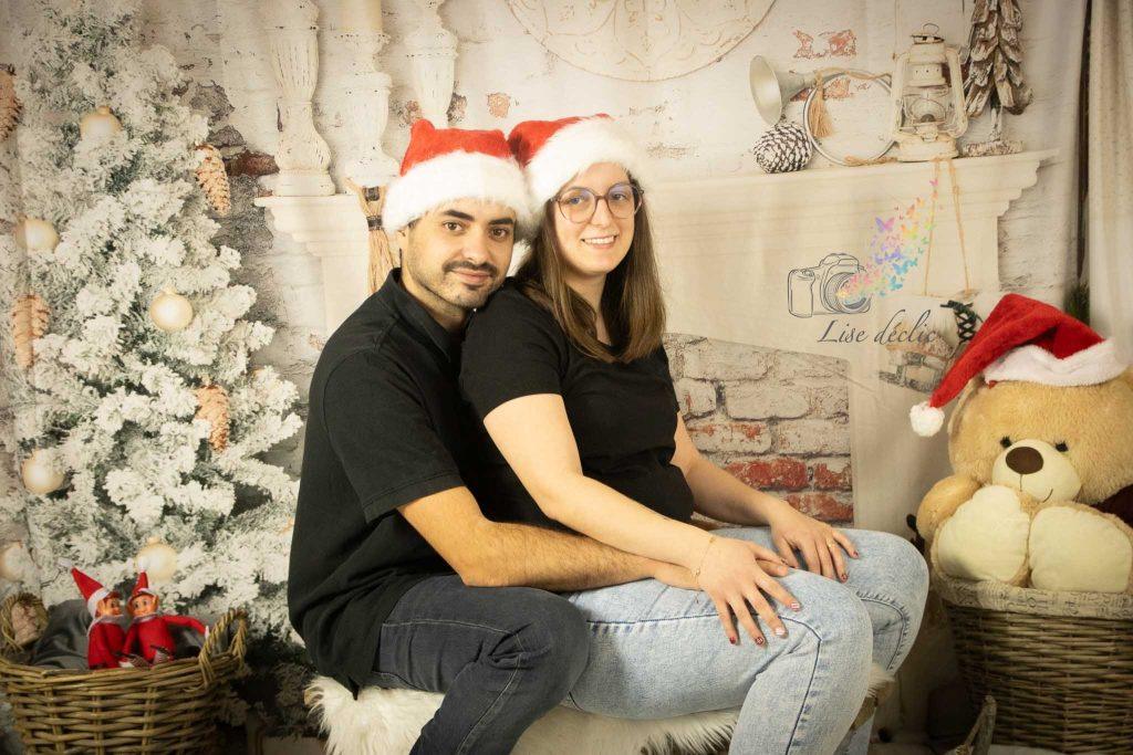 Couple assis devant un décor de Noël avec sapins enneigés et bonnets de Père Noël lors d’une mini séance photo à thème