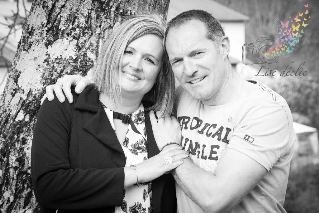 Couple souriant enlacé contre un arbre lors d’une séance photo authentique en extérieur en noir et blanc