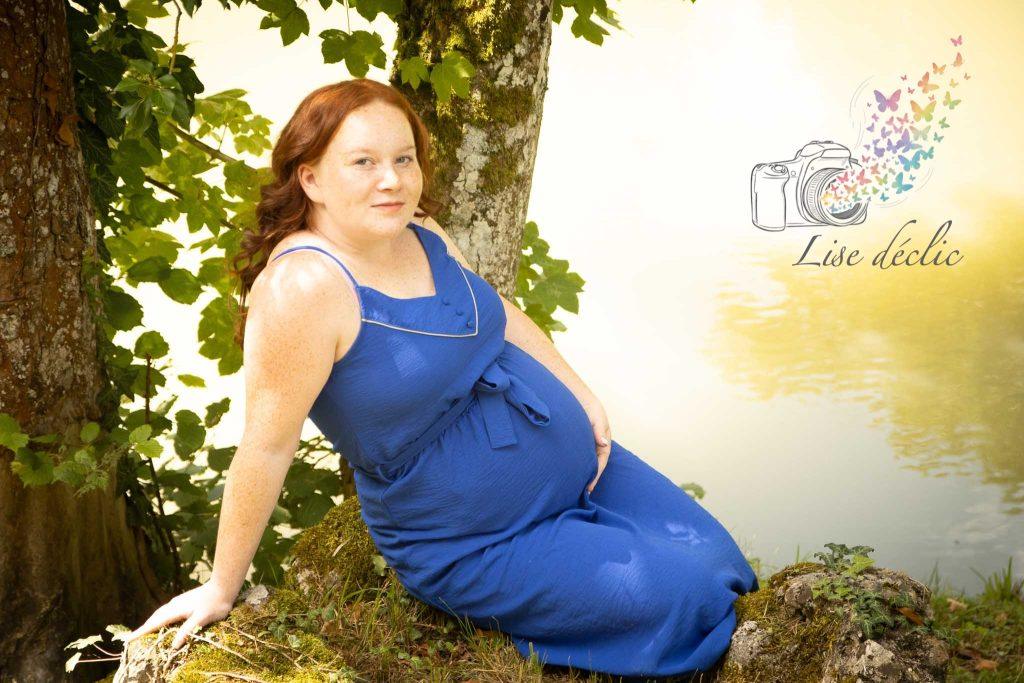 Femme enceinte assise près d’un arbre au bord de l’eau lors d’une séance photo grossesse en extérieur