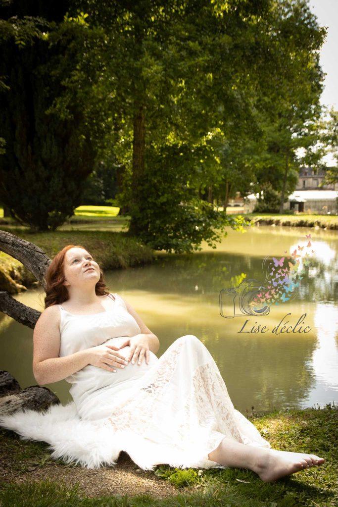 Femme enceinte assise au bord d’un étang sous un arbre lors d’une séance photo grossesse naturelle