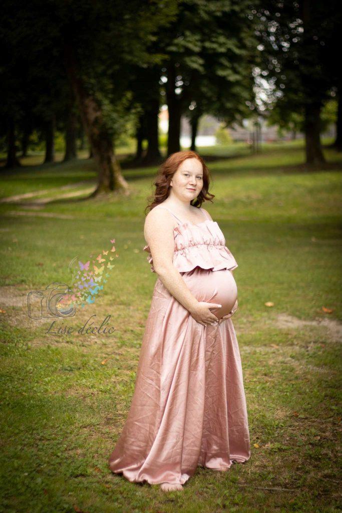 Femme enceinte debout dans un parc arboré portant une robe longue rose poudré lors d’une séance photo grossesse