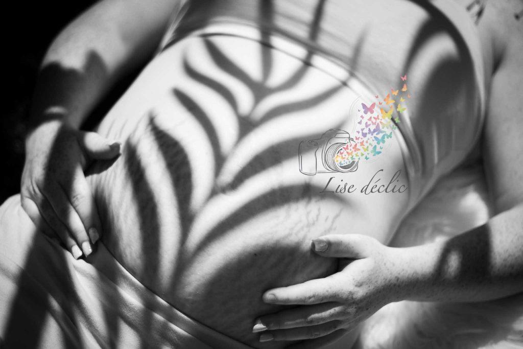 Gros plan en noir et blanc d’un ventre de femme enceinte avec jeu d’ombres naturelles lors d’une séance photo grossesse
